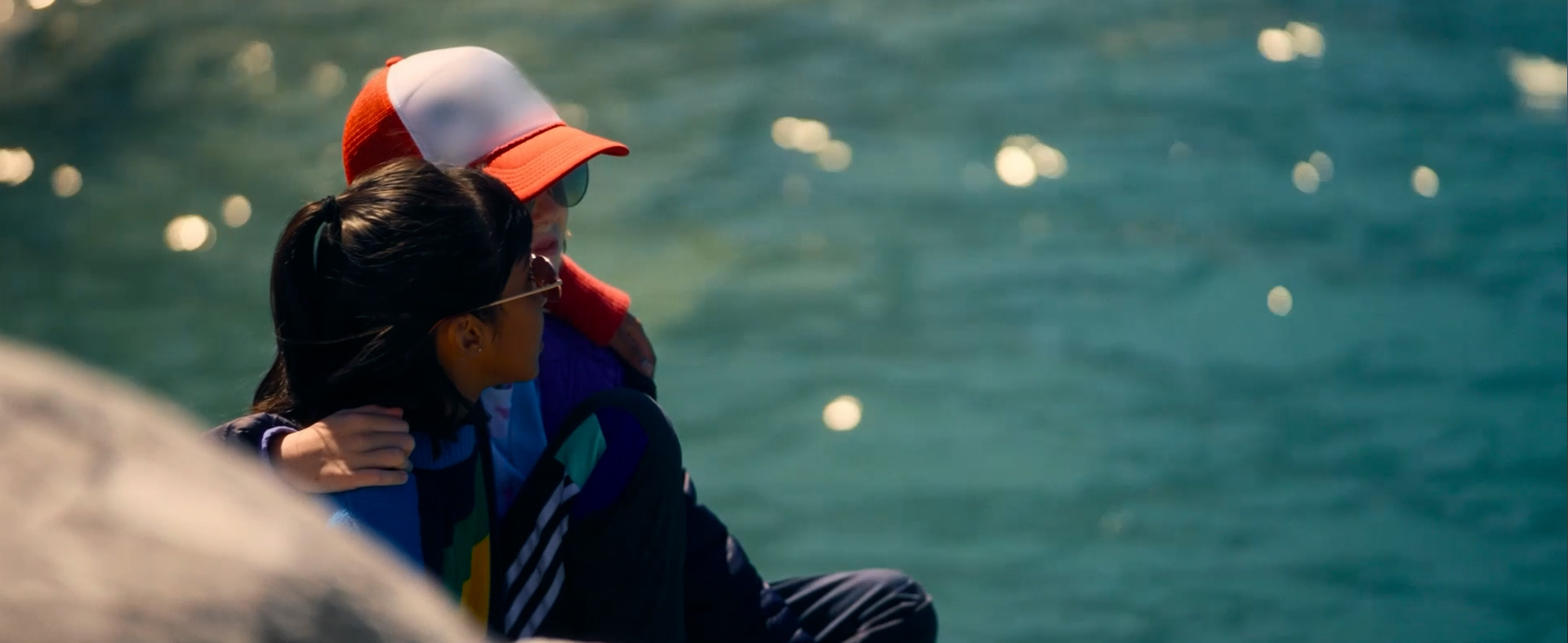 Zwei junge Personen sitzen im Freien am Wasser und umarmen sich. 