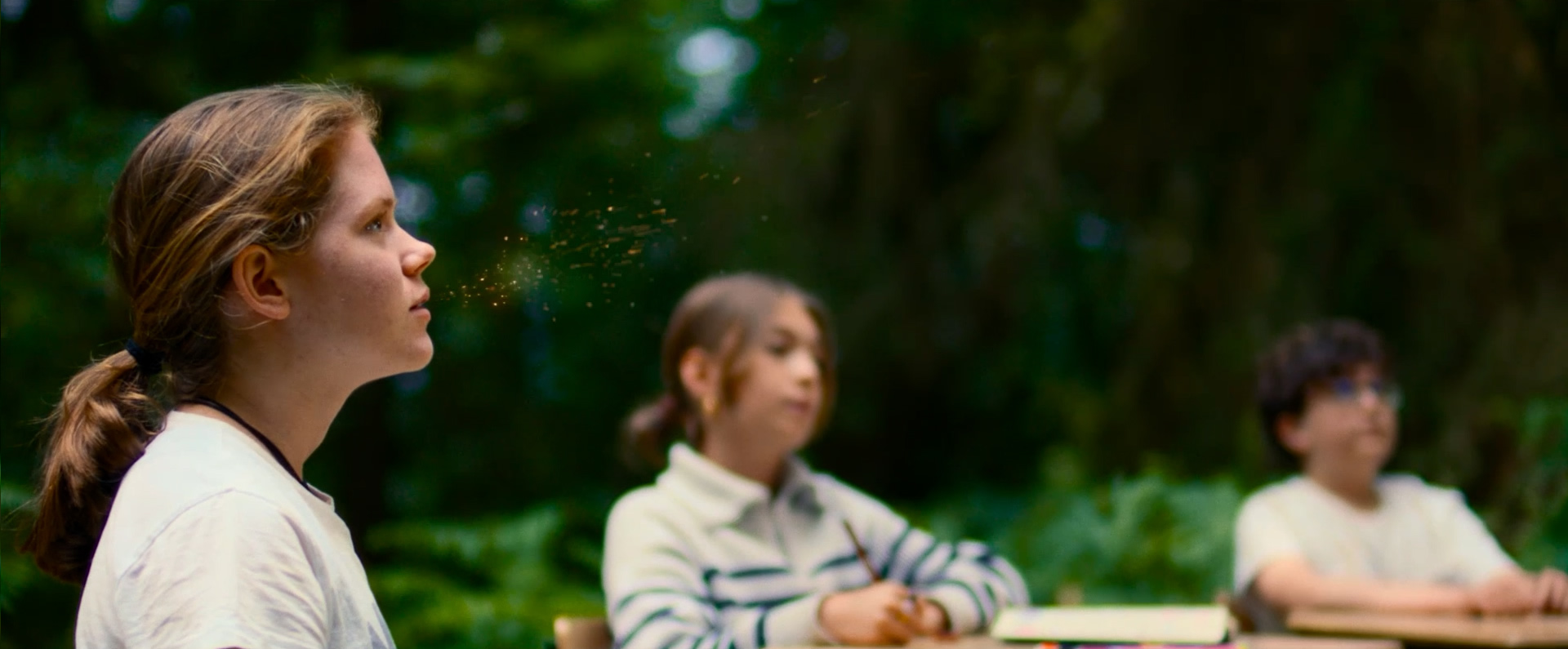 Drei Kinder sitzen an einem Tisch im Wald. Das Mädchen Skye im Vordergrund schaut seitlich nach oben, zwei weitere Kinder sind im Hintergrund zu sehen. Skye atmet goldene Partikelchen aus.