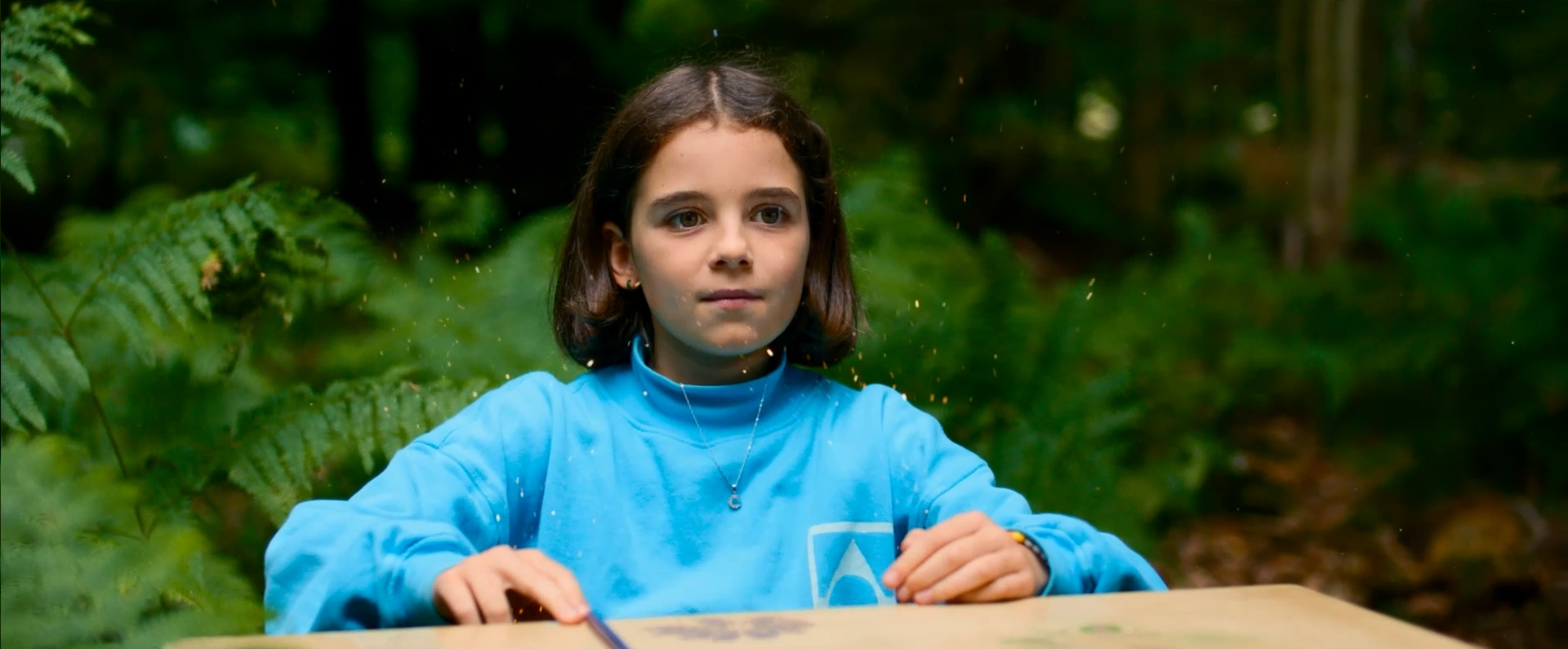 Filmstill CC im Wald: Das Mädchen Clemence mit schulterlangen dunklen Haaren und blauem Oberteil sitzt an einem Tisch im Wald und blickt in die Kamera. Im Hintergrund sind Farne und Bäume zu sehen. Vor ihrem Gesicht schweben goldene Punkte in der Luft.
