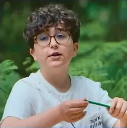 : Porträt eines Jungen mit lockigen dunklen Haaren und runder Brille. Er trägt ein weißes T-Shirt und hält einen Stift in den Händen. Im Hintergrund ist ein Wald zu sehen.