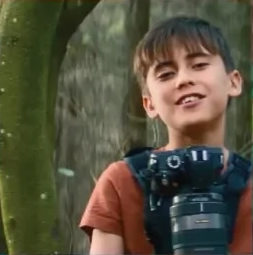 Porträt eines Jungen mit kurzen dunklen Haaren und braunem T-Shirt. Er trägt eine Kamera um den Hals und lächelt. Im Hintergrund ist ein Wald zu sehen.