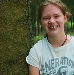 Porträt eines Mädchens mit rötlich-blonden Haaren und weißem T-Shirt. Sie lehnt an einem Baum und lächelt. Im Hintergrund ist ein Wald zu sehen.