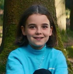 Porträt eines Mädchens mit schulterlangen dunklen Haaren und blauem Oberteil. Sie lächelt in die Kamera. Im Hintergrund ist ein Wald zu sehen.