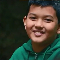 Porträt eines Jungen mit kurzen schwarzen Haaren und einem grünen Kapuzenpulli. Er lächelt in die Kamera vor einem dunklen Hintergrund.