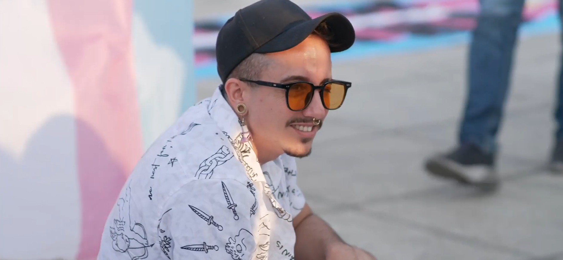 Nahaufnahme von Kani mit weißem Hemd, Base-Cap und Sonnenbrille. Im Hintergrund sind Transflaggen in Rosa, Hellblau und Weiß zu sehen.