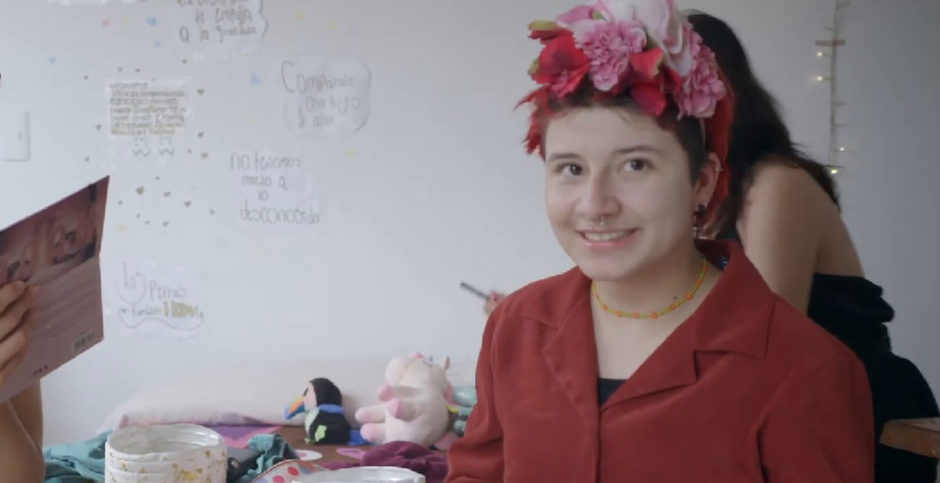 Oliver lächelt in die Kamera und trägt eine rote Bluse sowie rote Plastikblumen im Haar. Im Hintergrund schminken sich Karla und Valentina auf dem Tisch liegt ein kleines rosa Stoff-Tier.