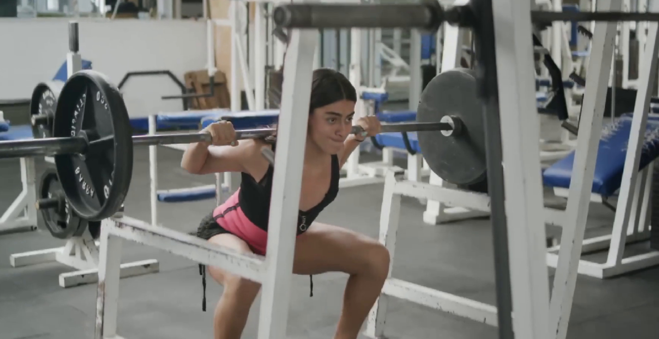 Karla führt in einem Fitnessstudio mit einer Langhantel auf den Schultern eine Kniebeuge aus und lächelt dabei angestrengt.