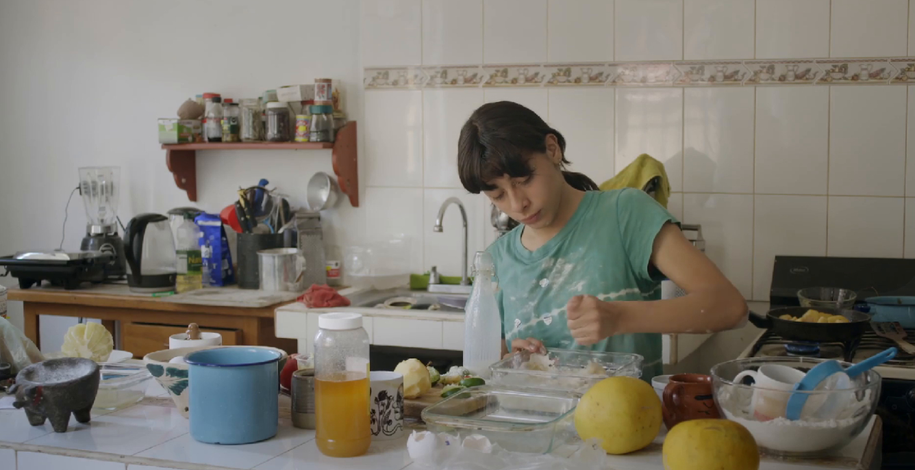 Karla füllt in der Küche ihrer Eltern Mehl mit der Hand in eine Auflaufform. Küchengeräte, Schüsseln und Lebensmittel stehen auf der mit Mehl bestäubten Arbeitsfläche.