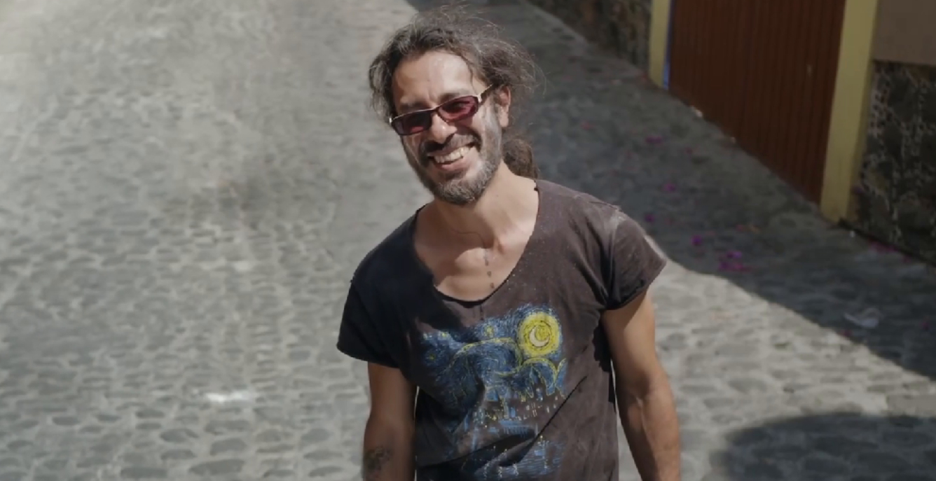 Karlas Vater lacht in die Kamera und steht auf einer Straße in Tepoztlán. Er trägt ein gemustertes braunes T-Shirt, eine Sonnenbrille und einen Bart, seine langen Haare sind zum Zopf gebunden.