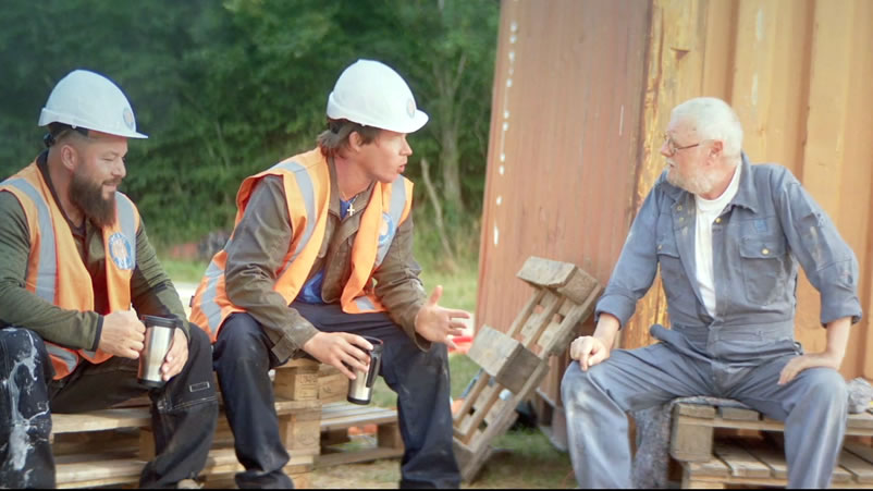 Filmbild: Zwei Bauarbeiter und ein älterer Mann sitzen zusammen und unterhalten sich.